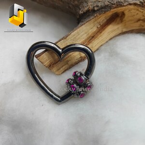 May include: A black heart-shaped charm with a small cluster of pink gemstones in the center. The charm is made of metal and has a shiny finish.