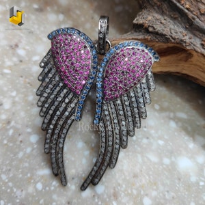 Puede incluir: Un colgante de plata con un par de alas con piedras preciosas rosas y azules. Las alas están engastadas con pequeñas piedras brillantes y tienen un diseño delicado e intrincado.