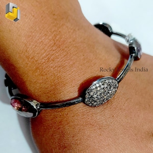 May include: A silver bangle bracelet featuring a large oval-shaped gemstone set with small diamonds. The bracelet also includes smaller gemstones in various colours. The bracelet is worn on a person's wrist. "Rocky Jewels India" is inscribed on the bracelet.