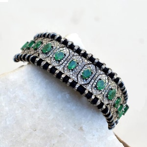Puede incluir: Un brazalete de plata con esmeraldas ovaladas y pequeños diamantes engastados en un diseño geométrico. El brazalete tiene un detalle de cordón negro entre las secciones plateadas. El brazalete se muestra sobre una superficie blanca.