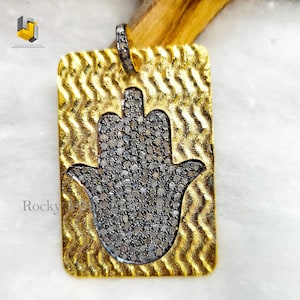 Può includere: Un ciondolo in oro con un design a mano di hamsa, incastonato con piccoli diamanti. Il ciondolo ha una superficie testurizzata e ondulata.