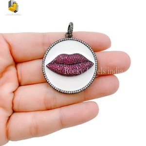 May include: A silver pendant with a white enamel circle and a red rhinestone lip design. The pendant is surrounded by smaller rhinestones.