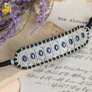 Può includere: Un braccialetto in argento e nero con pietre preziose con zaffiri blu ovali e piccoli diamanti bianchi. Il braccialetto è montato su un cordoncino nero e poggia su uno sfondo di carta vintage.