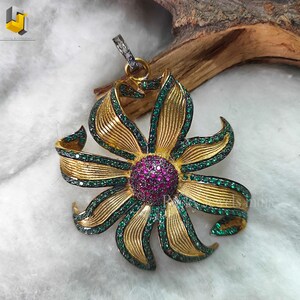 May include: A gold pendant in the shape of a flower with green and red gemstones. The flower has a central red gemstone surrounded by smaller green gemstones. The pendant is on a white background.