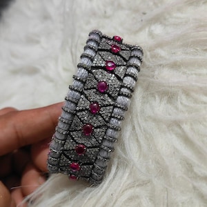 May include: A silver-toned bracelet with a geometric design, featuring small, sparkling accents and vibrant red gemstones. The bracelet is rectangular and appears to be made of metal and fabric.