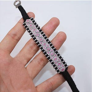 May include: A black bracelet with a decorative design featuring pink and clear gemstones. The bracelet has a woven black band and a silver clasp. The gemstones are arranged in a repeating pattern.