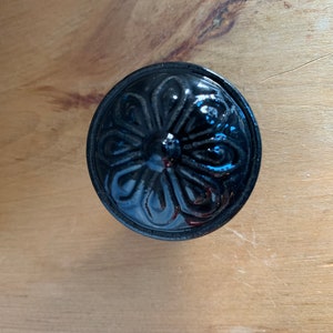 May include: A black, decorative lid with a floral design. The lid is round and has a glossy finish. The image is taken from a top-down perspective, showcasing the intricate details of the embossed pattern.
