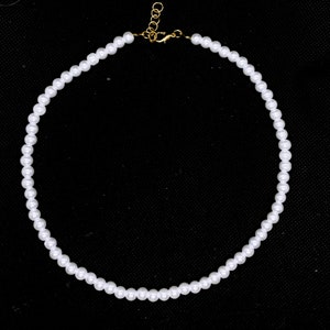 May include: A delicate pearl necklace with small, round, white beads. The necklace has a gold-colored clasp and extender chain. The pearls are strung together to form a complete circle, set against a black background.