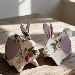 May include: Two fabric rabbit figurines featuring a cream-colored base with a floral pattern of pink roses and green leaves. Each rabbit has solid lavender-colored ears and side panels, with a small white pom-pom tail attached to the back.