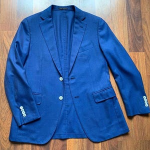 Könnte beinhalten: Ein leuchtend blauer Blazer mit klassischem Zwei-Knopf-Verschluss und fallendem Revers. Die Jacke hat zwei Vordertaschen und dekorative Knöpfe an den Ärmeln. Der Blazer ist aus einem strukturierten Stoff gefertigt.