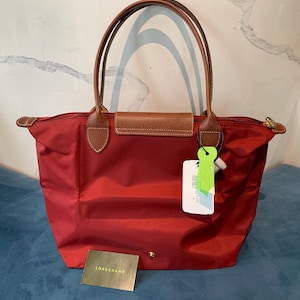 May include: A red tote bag with brown leather handles and accents. The bag has a rectangular shape with a zipper closure and a flap with a snap closure. A small gold-colored card and a tag are attached to the bag.