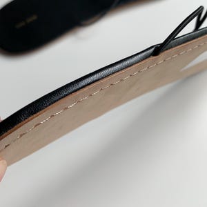 May include: A pair of black flat sandals featuring thin, delicate straps and a minimalist design. The sole has a smooth, light-coloured underside with visible stitching along the edge, while the top surface is black with a subtle brand logo.