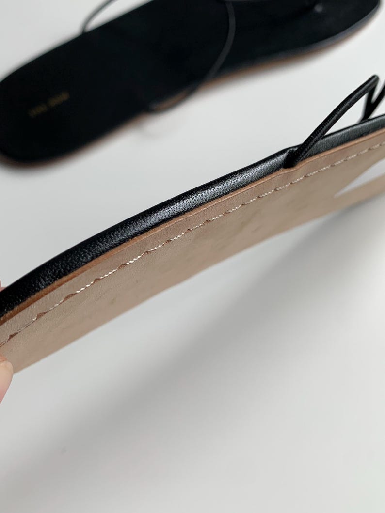 May include: A pair of black flat sandals featuring thin, delicate straps and a minimalist design. The sole has a smooth, light-coloured underside with visible stitching along the edge, while the top surface is black with a subtle brand logo.