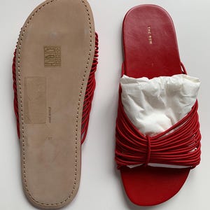 May include: A pair of red slide sandals featuring a multi-strand corded strap design. One sandal shows the beige leather sole with size 37 and Made in Italy text, while the other displays the red leather footbed with The Row logo in gold.