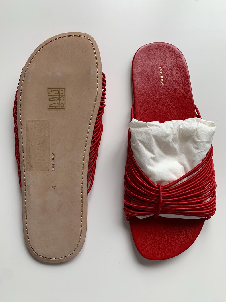 May include: A pair of red slide sandals featuring a multi-strand corded strap design. One sandal shows the beige leather sole with size 37 and Made in Italy text, while the other displays the red leather footbed with The Row logo in gold.