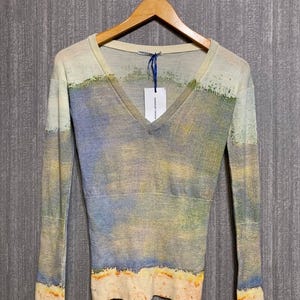 Könnte beinhalten: Ein langärmliger V-Ausschnitt-Pullover mit Landschaftsdesign. Der Pullover hat eine Farbpalette aus Blau, Gelb und Beige mit strukturiertem Aussehen. Das Kleidungsstück hängt an einem Holzbügel.