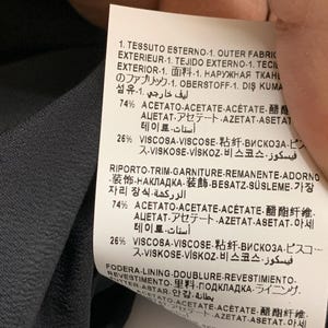 K&ouml;nnte beinhalten: Nahaufnahme eines Stoffetiketts mit Text in mehreren Sprachen. Das Etikett listet die prozentuale Zusammensetzung des Stoffes auf, einschlie&szlig;lich Acetat und Viskose. Der Hintergrund ist ein dunkelgrauer Stoff.