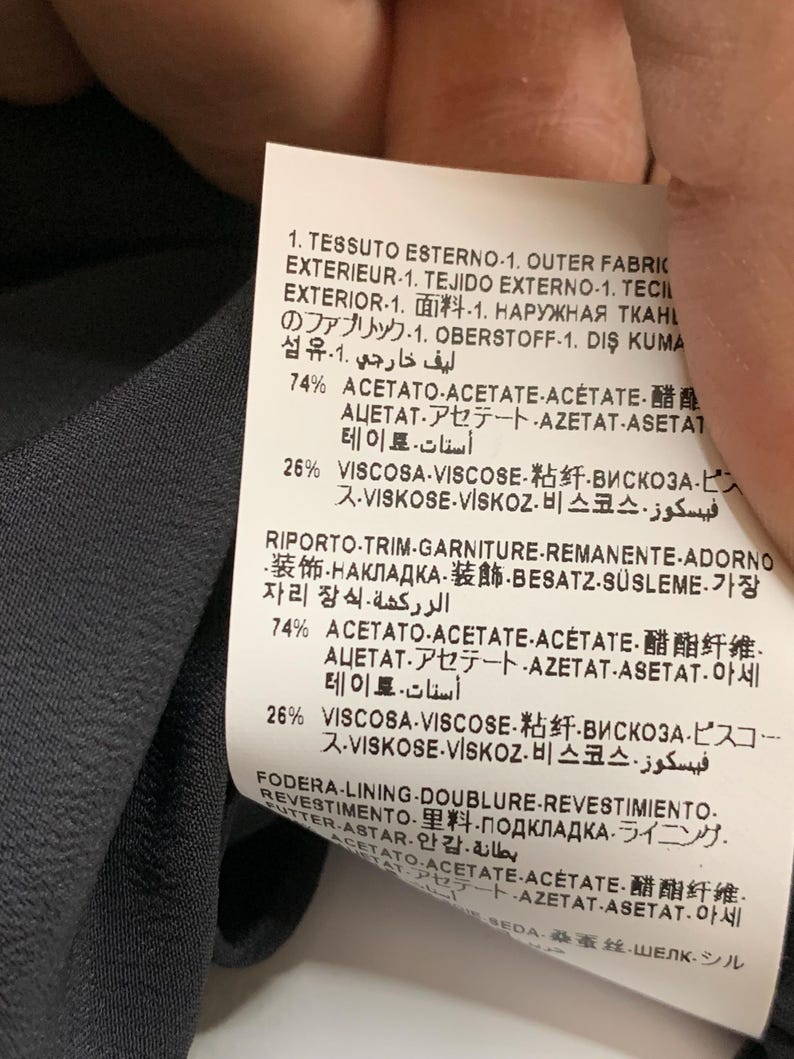 K&ouml;nnte beinhalten: Nahaufnahme eines Stoffetiketts mit Text in mehreren Sprachen. Das Etikett listet die prozentuale Zusammensetzung des Stoffes auf, einschlie&szlig;lich Acetat und Viskose. Der Hintergrund ist ein dunkelgrauer Stoff.