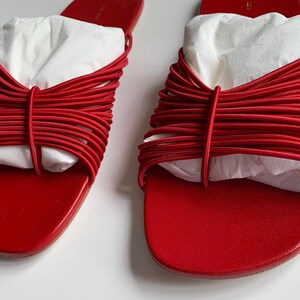 May include: A pair of bright red slide sandals featuring a series of thin, horizontal elastic cords across the top strap. The sandals have a smooth, flat footbed and are stuffed with white paper inserts to maintain their shape and structure.