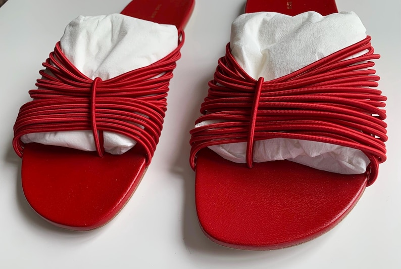 May include: A pair of bright red slide sandals featuring a series of thin, horizontal elastic cords across the top strap. The sandals have a smooth, flat footbed and are stuffed with white paper inserts to maintain their shape and structure.