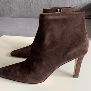 May include: A pair of dark brown suede ankle boots featuring a pointed toe, a slim stiletto heel, and a side zip closure. The boots have a smooth, matte texture and a clean, structured silhouette that reaches just above the ankle bone.