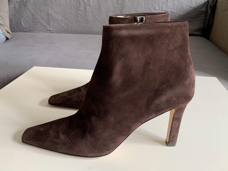 May include: A pair of dark brown suede ankle boots featuring a pointed toe, a slim stiletto heel, and a side zip closure. The boots have a smooth, matte texture and a clean, structured silhouette that reaches just above the ankle bone.