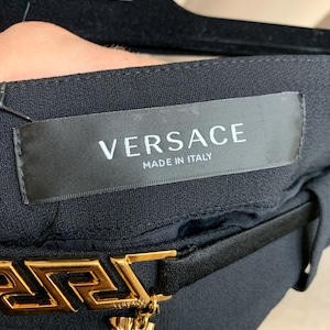 K&ouml;nnte beinhalten: Schwarzer Stoff mit goldfarbener Versace-G&uuml;rtelschnalle. Auf dem Etikett steht "VERSACE MADE IN ITALY". Das Versace-Logo ist auf dem Etikett und der G&uuml;rtelschnalle zu sehen. Die G&uuml;rtelschnalle hat ein griechisches Schl&uuml;sseldesign.