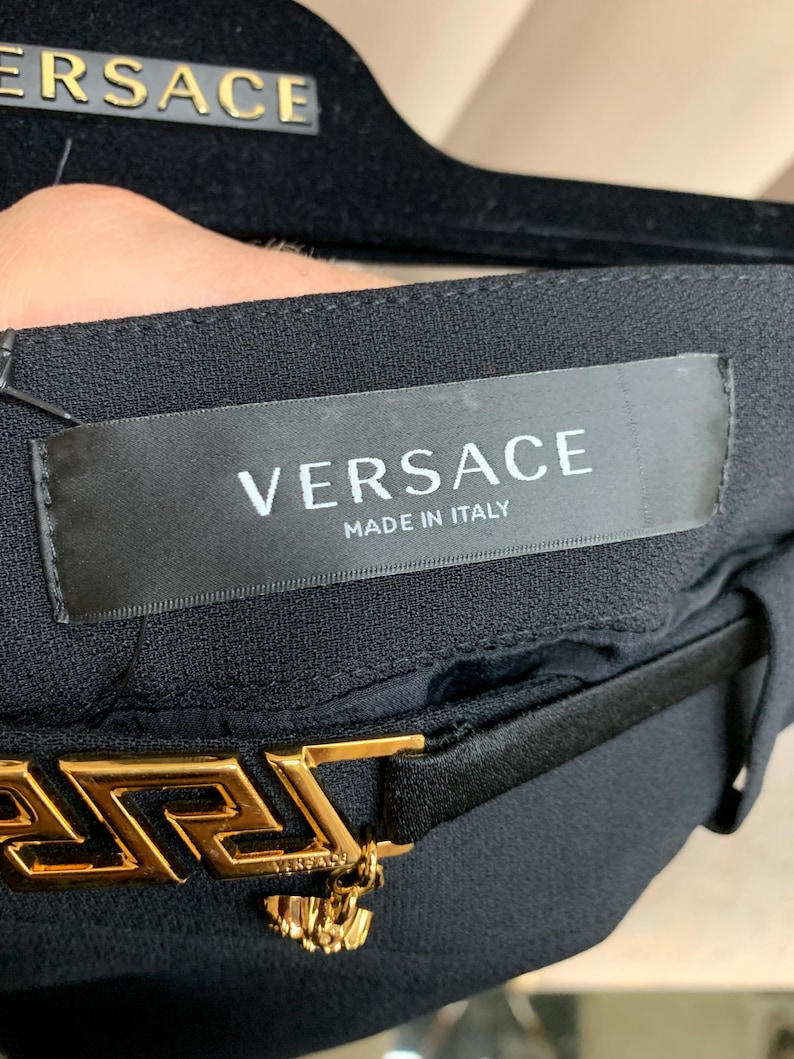 K&ouml;nnte beinhalten: Schwarzer Stoff mit goldfarbener Versace-G&uuml;rtelschnalle. Auf dem Etikett steht "VERSACE MADE IN ITALY". Das Versace-Logo ist auf dem Etikett und der G&uuml;rtelschnalle zu sehen. Die G&uuml;rtelschnalle hat ein griechisches Schl&uuml;sseldesign.