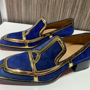 May include: A pair of blue suede loafers with gold trim. The shoes have a square toe and a small black heel. The gold trim outlines the vamp and sides of the shoe, creating a geometric design. The shoes are displayed on a white surface.