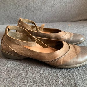 May include: A pair of brown leather Mary Jane style shoes with ankle straps. The shoes have a rounded toe and a low heel. The leather has a distressed look. The shoes are sitting on a gray surface.
