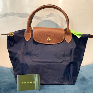 Op de afbeelding: Een marineblauwe handtas met bruine leren handvatten en een klepsluiting. De tas heeft een goudkleurige sluiting en een klein groen label. De merknaam "LONGCHAMP" staat op een groene kaart.