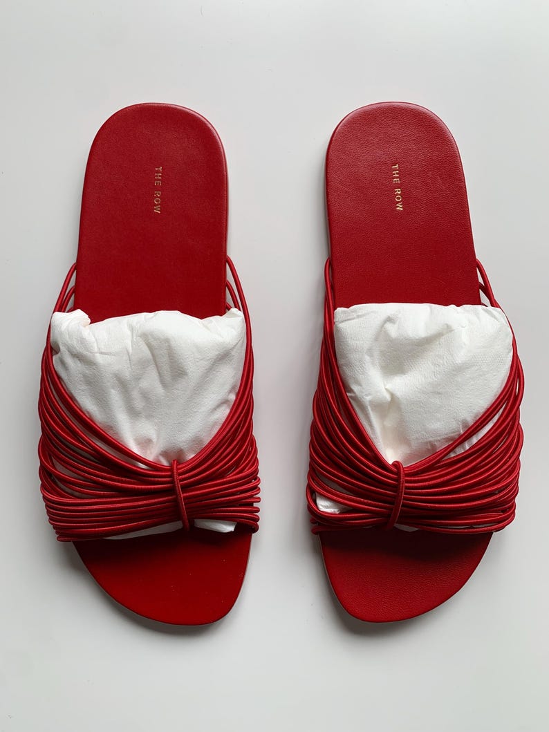 May include: A pair of bright red slide sandals featuring a flat sole and a wide strap composed of multiple thin, parallel red cords gathered in the center. The brand name THE ROW is printed in gold on the heel area of the red footbed.