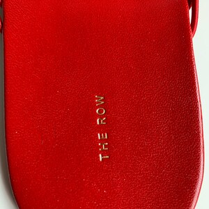 May include: A bright red sandal footbed with a textured surface and the text THE ROW embossed in gold lettering in the center. The sandal features a smooth, rounded heel area and a thin red strap visible at the top edge of the footwear design.