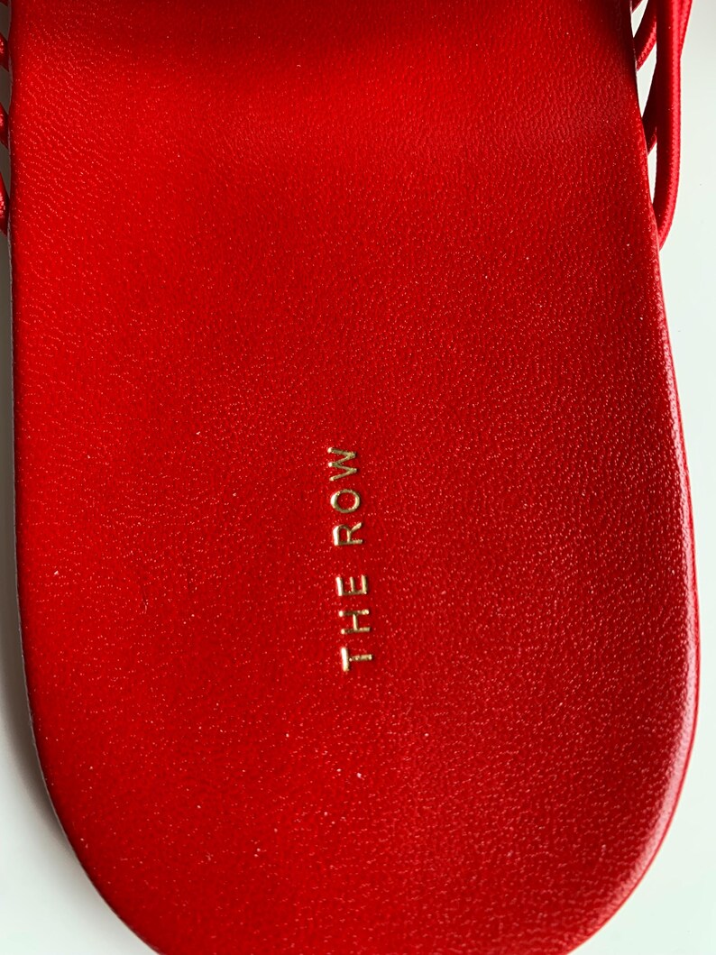 May include: A bright red sandal footbed with a textured surface and the text THE ROW embossed in gold lettering in the center. The sandal features a smooth, rounded heel area and a thin red strap visible at the top edge of the footwear design.