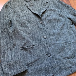 Pode incluir: Um blazer cinza em espinha de peixe com gola entalhada, fecho de três botões e dois bolsos frontais. O blazer tem um design clássico e é feito de um tecido texturizado. A etiqueta diz "ORKA".