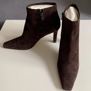 May include: A pair of dark brown ankle boots with a pointed toe and a slim heel. The boots are crafted from a soft, suede-like material and feature a side zip closure. White paper inserts are visible inside the top opening of each boot.