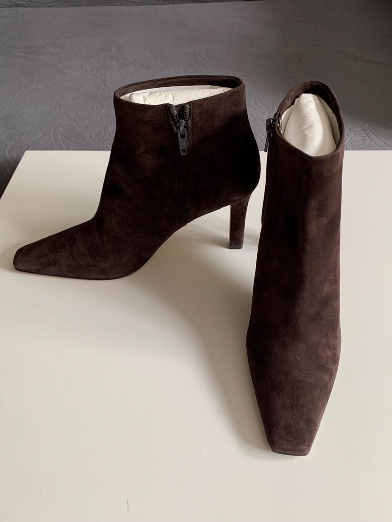May include: A pair of dark brown ankle boots with a pointed toe and a slim heel. The boots are crafted from a soft, suede-like material and feature a side zip closure. White paper inserts are visible inside the top opening of each boot.