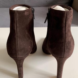May include: A pair of dark brown suede ankle boots with a high heel and a side zip closure. The boots feature a smooth, matte texture and a structured silhouette, showing the back view of the footwear with a clean, solid colour finish.
