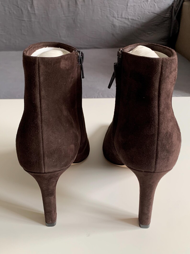May include: A pair of dark brown suede ankle boots with a high heel and a side zip closure. The boots feature a smooth, matte texture and a structured silhouette, showing the back view of the footwear with a clean, solid colour finish.