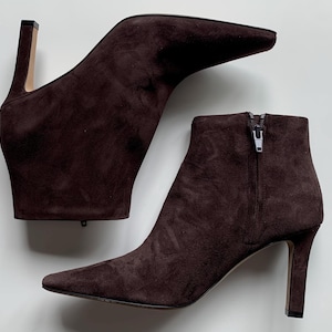 May include: A pair of dark brown suede ankle boots featuring a pointed toe, a slim high heel, and a side zip closure. The boots have a smooth, matte texture and a structured silhouette, with one boot positioned horizontally above the other.