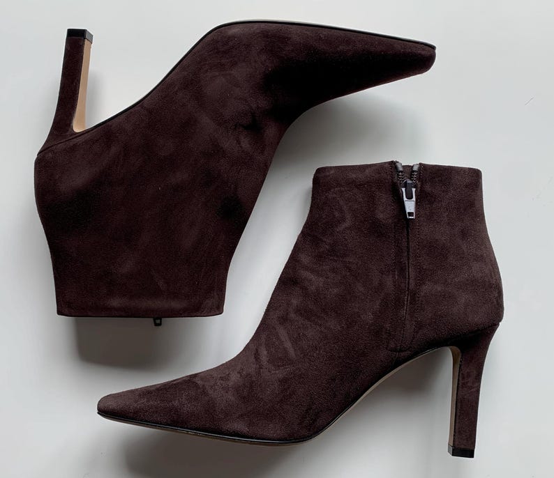 May include: A pair of dark brown suede ankle boots featuring a pointed toe, a slim high heel, and a side zip closure. The boots have a smooth, matte texture and a structured silhouette, with one boot positioned horizontally above the other.