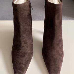 May include: A pair of dark brown suede ankle boots with a square toe design. Each boot features a side zip fastening and a slim heel. The interior of the boots is filled with white paper stuffing to maintain their upright shape and structure.