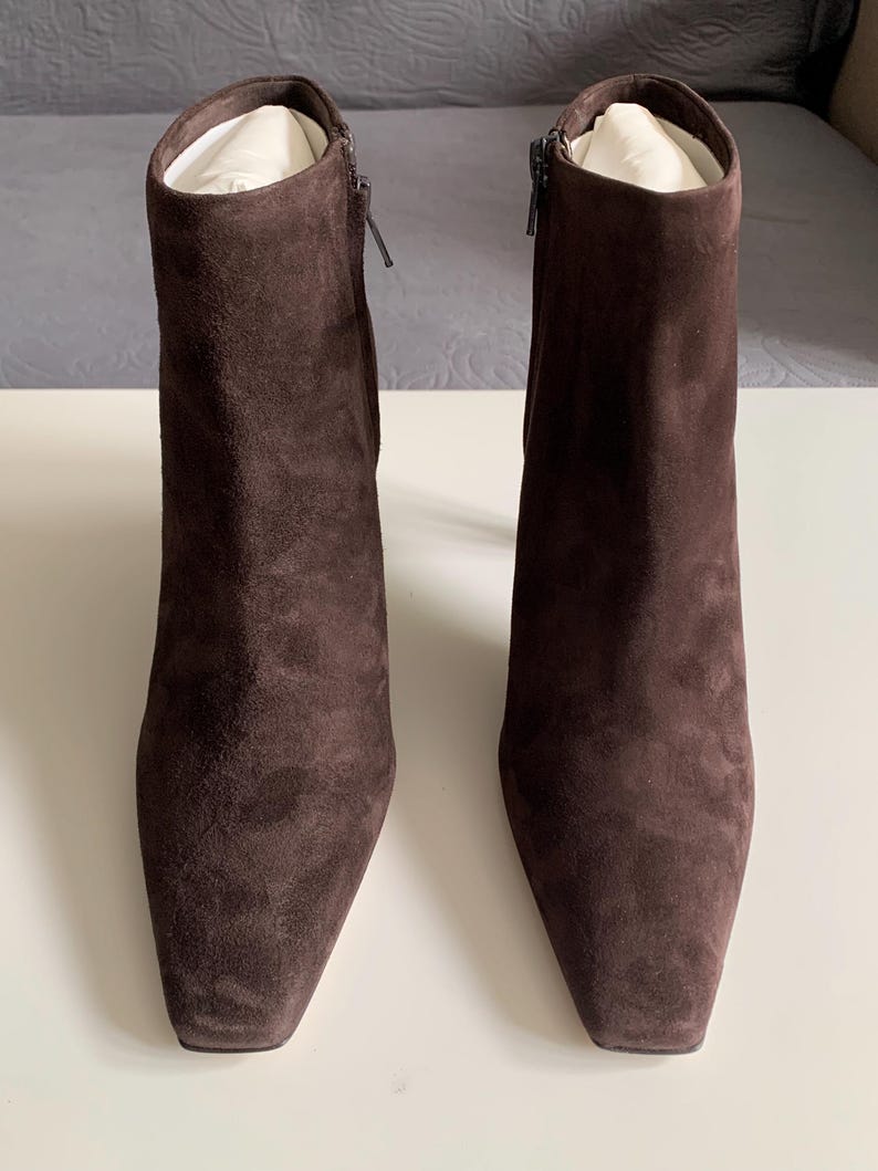 May include: A pair of dark brown suede ankle boots with a square toe design. Each boot features a side zip fastening and a slim heel. The interior of the boots is filled with white paper stuffing to maintain their upright shape and structure.