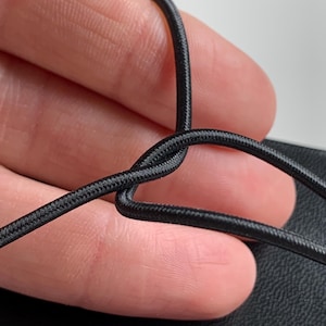 May include: A thin, black, flexible elastic cord with a woven, textured surface. The cord is bent into a loop shape, showing its round profile and consistent dark black colour throughout the entire length of the material visible in the frame.