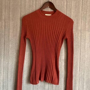 Könnte beinhalten: Ein rostfarbener Rippstrickpullover mit langen Ärmeln und Rundhalsausschnitt. Der Pullover hängt an einem Holzkleiderbügel vor einer strukturierten, neutral getönten Wand. Die Rippstruktur verläuft vertikal.