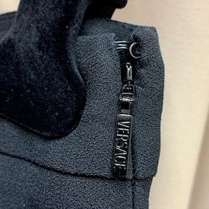 K&ouml;nnte beinhalten: Nahaufnahme eines schwarzen Kleidungsst&uuml;cks mit Samtdetail und Rei&szlig;verschluss. Der Rei&szlig;verschlussanh&auml;nger ist rechteckig und mit dem Wort "VERSACE" gepr&auml;gt. Der Stoff hat ein strukturiertes Aussehen.