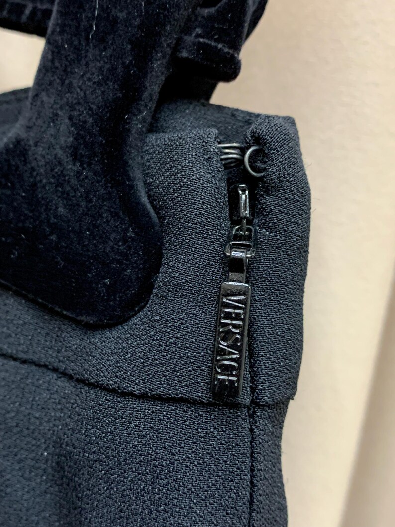 K&ouml;nnte beinhalten: Nahaufnahme eines schwarzen Kleidungsst&uuml;cks mit Samtdetail und Rei&szlig;verschluss. Der Rei&szlig;verschlussanh&auml;nger ist rechteckig und mit dem Wort "VERSACE" gepr&auml;gt. Der Stoff hat ein strukturiertes Aussehen.