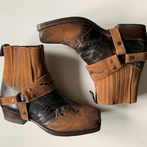 Könnte beinhalten: Ein Paar braune und schwarze Leder-Ankle Boots. Die Stiefel haben ein Used-Finish, dekorative Nähte und Lederriemen mit Metallbeschlägen. Sie haben einen Blockabsatz und eine spitze Zehenpartie.