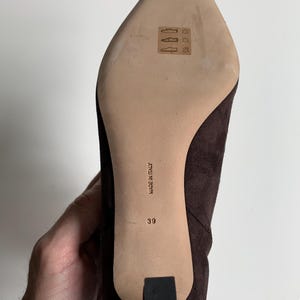 May include: A dark brown suede shoe with a pointed toe and a heel. The light-coloured sole features a stamped leather icon, the text "MADE IN ITALY," and the size number "39" printed in black. The shoe has a textured finish and a black heel cap.