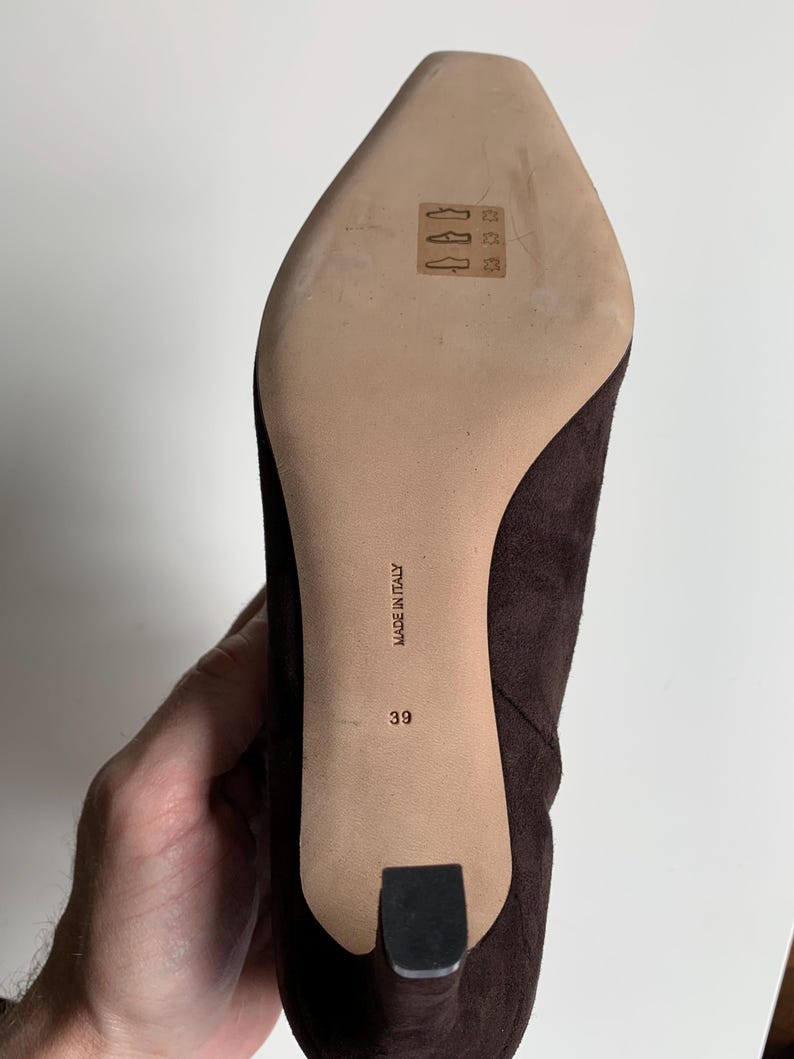 May include: A dark brown suede shoe with a pointed toe and a heel. The light-coloured sole features a stamped leather icon, the text "MADE IN ITALY," and the size number "39" printed in black. The shoe has a textured finish and a black heel cap.